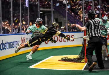 A lacrosse player dives towards the goal while another player and a referee are nearby, amidst a cheering crowd.