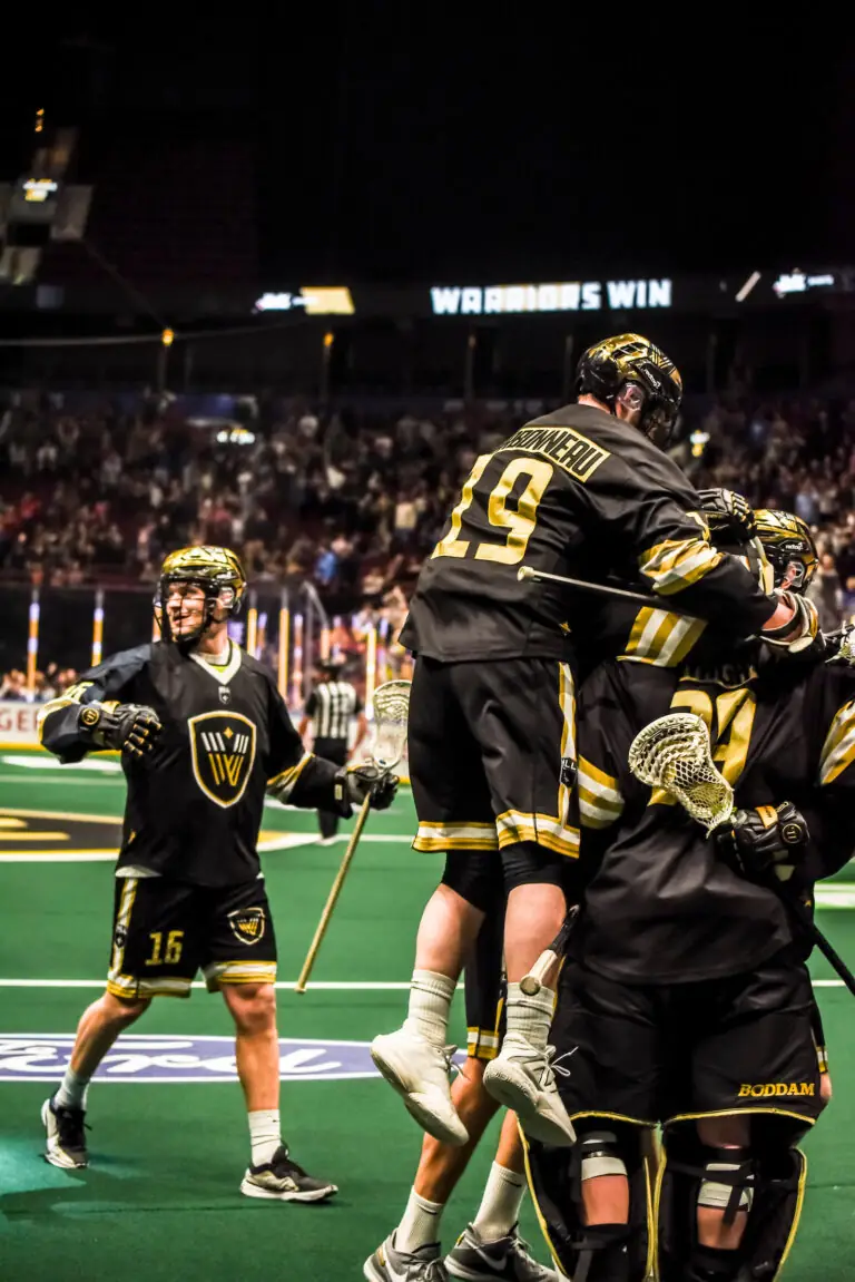 Lacrosse players celebrate a goal, with one player lifted in joy, on a green field.