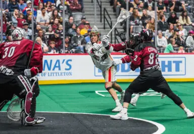Lacrosse player shooting the ball while two defenders and a goalie are positioned nearby in a stadium filled with spectators.