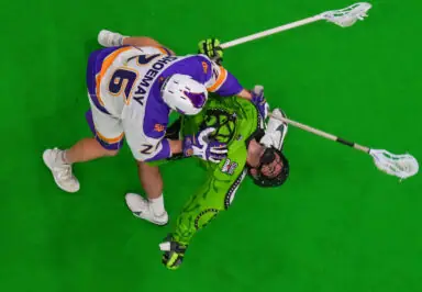 Two lacrosse players engage in a face-off on a bright green field. One player in a white and purple uniform exerts pressure on the other, who wears a green uniform.