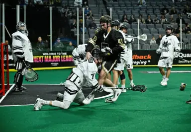 A lacrosse player in black jerseys engages with an opponent in white while a goalie watches from the net. Spectators are visible in the background.