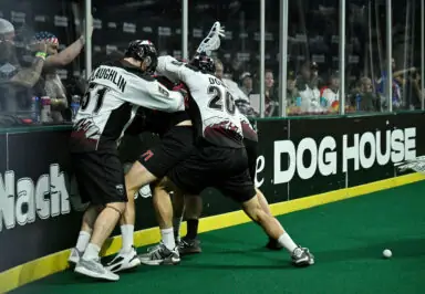 Lacrosse players engage in a physical struggle near the boards during a game, with spectators visible in the background.