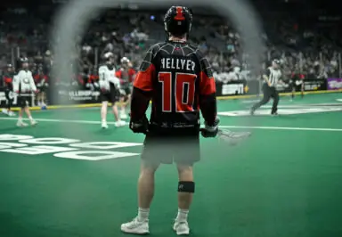 Player wearing a black and red lacrosse jersey with the name "HELLYER" and number 10, standing on the field during a game.