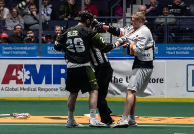 A lacrosse player in a black jersey labeled "BILES" grapples with an opposing player while a referee intervenes on the field.