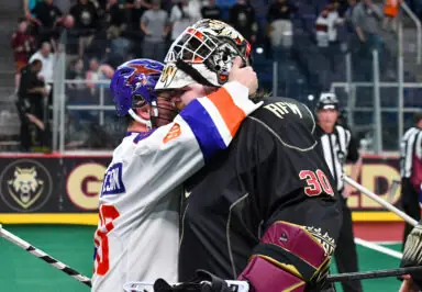 Two lacrosse players embrace after a match, one wearing a white jersey and the other in black goalie gear.