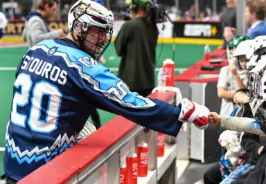 A lacrosse player in a blue jersey fist bumps a young fan in a helmet at the sidelines.
