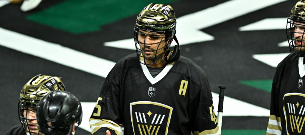 A lacrosse player in a black jersey with gold accents and the letter "A" on the chest talks with teammates on the field.