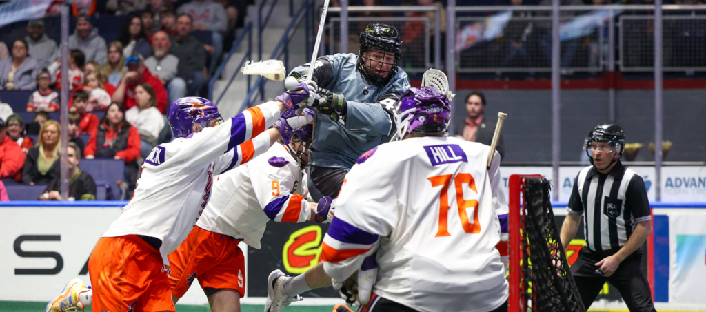 A lacrosse player in gray jumps over two opponents in orange and purple, while a referee watches the action near the goal.