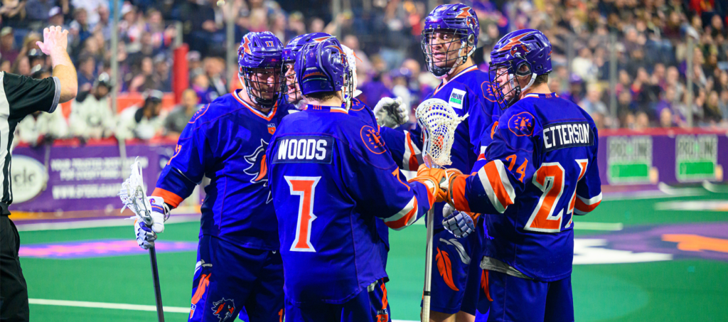 A group of lacrosse players in blue jerseys huddle together on the field, with a referee signaling nearby.