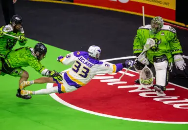 A lacrosse player in a white and purple uniform stretches to shoot while being defended by two players in green uniforms, with a goalie in green ready to block the shot.