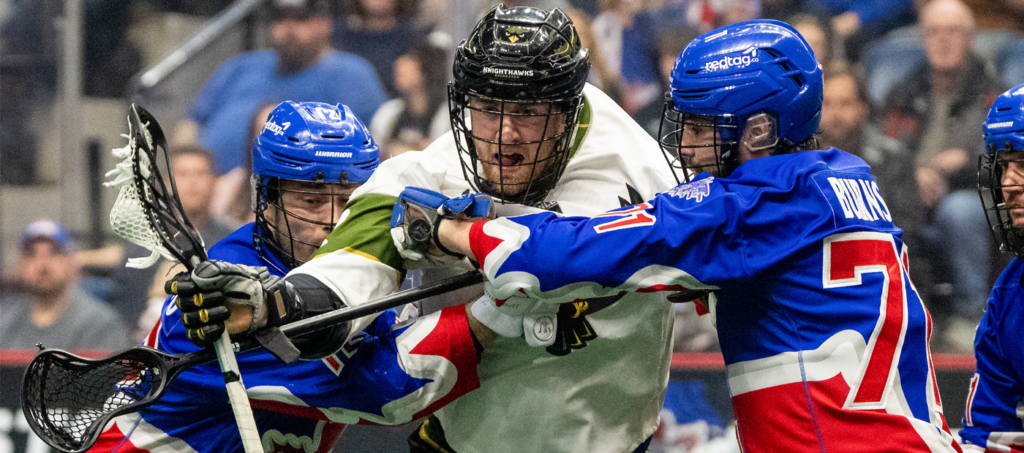 A lacrosse player in a white uniform is being challenged by two players in blue uniforms during a game.