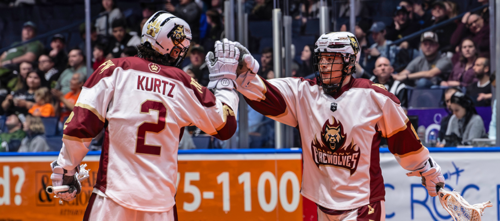 Two lacrosse players in white and maroon jerseys celebrate with a high-five during a game, with fans visible in the background.
