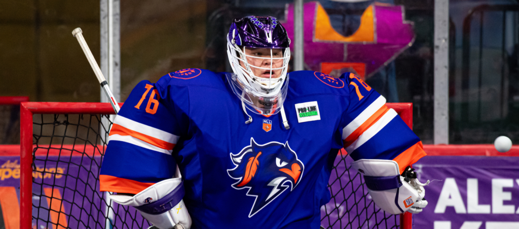 Goalkeeper in blue lacrosse uniform, standing in front of a goal net.