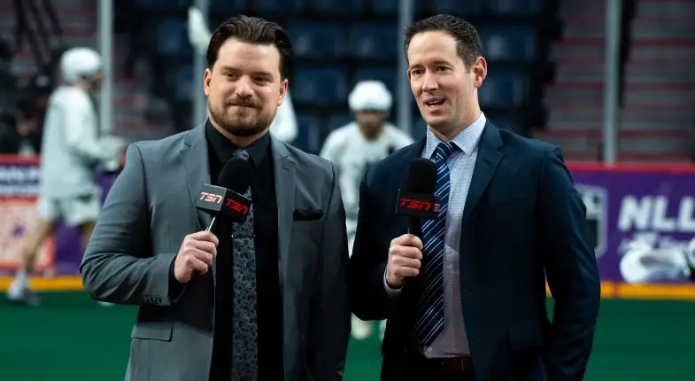 Two sports commentators in suits hold microphones while standing on a lacrosse field.
