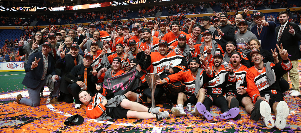 A group of lacrosse players and supporters wearing orange and black team jerseys celebrate with a trophy amidst confetti on the field.