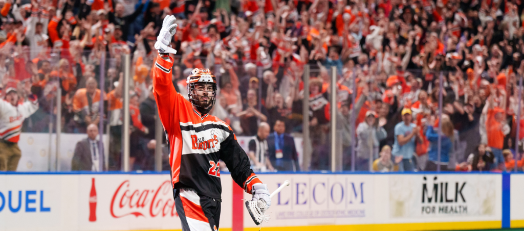 Lacrosse player in an orange jersey waves to a cheering crowd during a game.