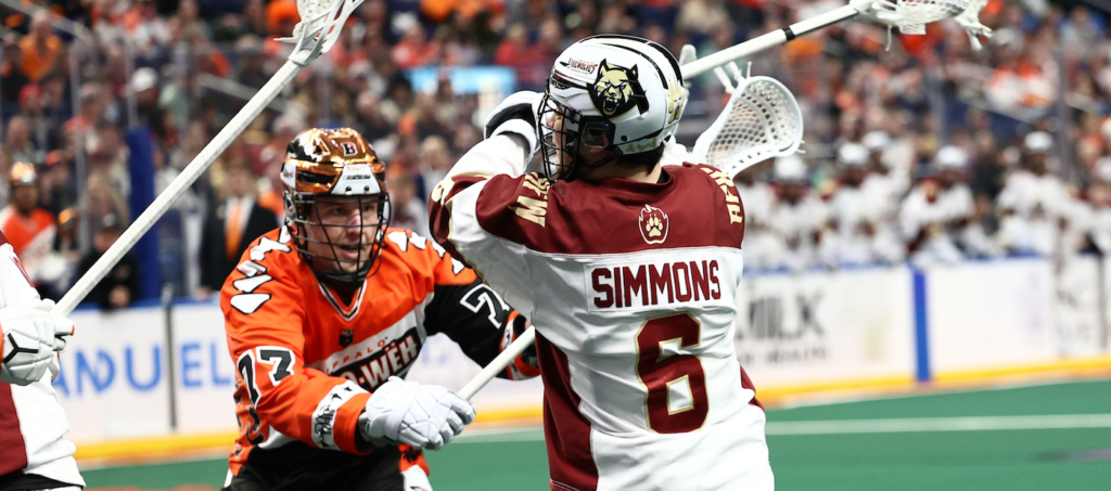 Two lacrosse players compete in a match, one wearing a white jersey with "SIMMONS" and the number 6, while the other wears an orange jersey.