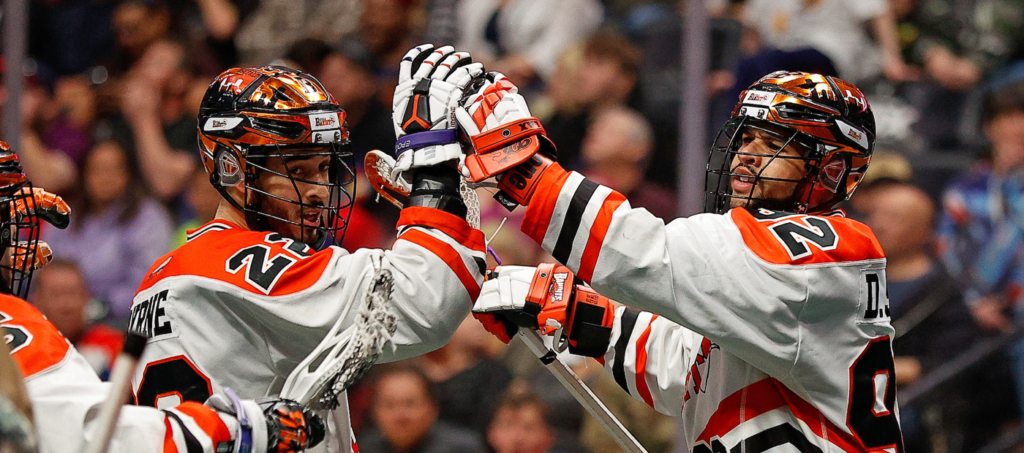 Two lacrosse players celebrate a goal, raising their sticks in a crowded arena.