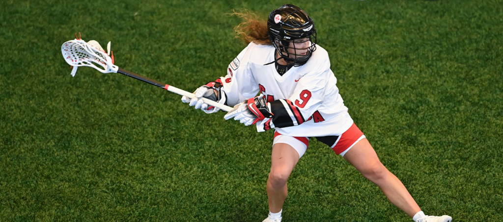 A female lacrosse player in a white jersey with red and black accents is in mid-action, preparing to pass or shoot the ball on a green turf field.