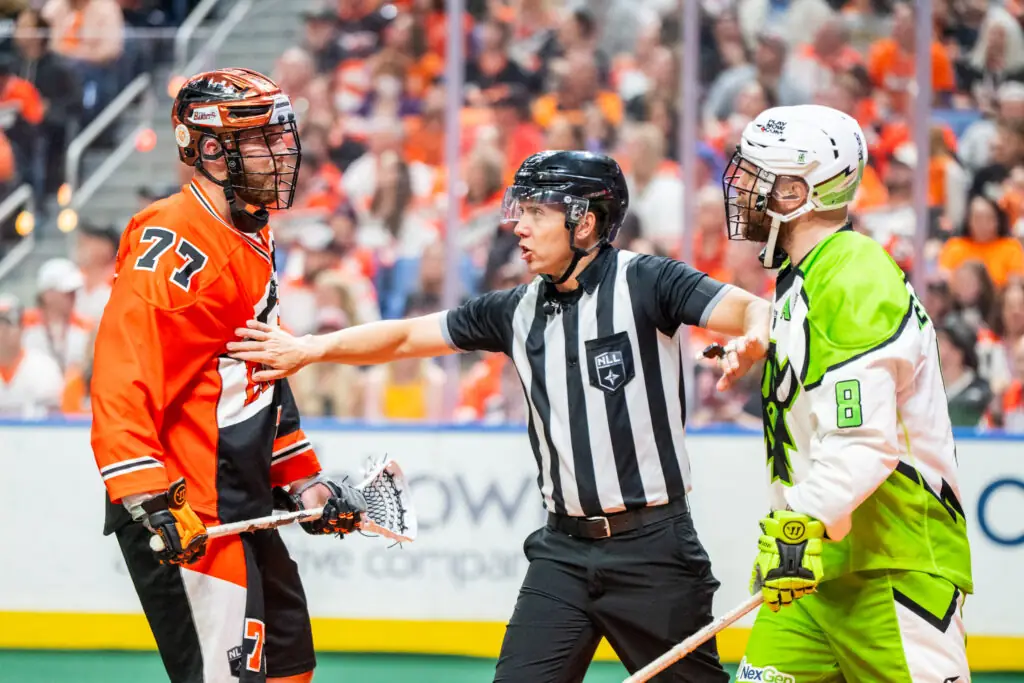 Referee separates two lacrosse players in orange and green uniforms
