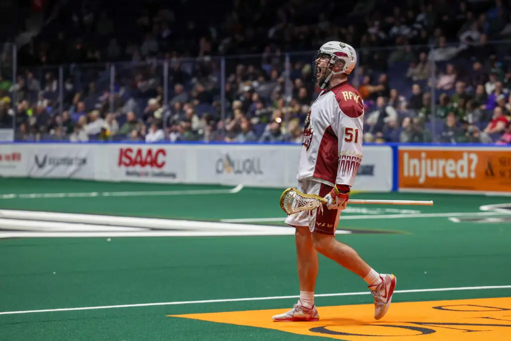 Lacrosse player wearing white and red uniform, holding a stick, walks across the field. Audience blurred in background.
