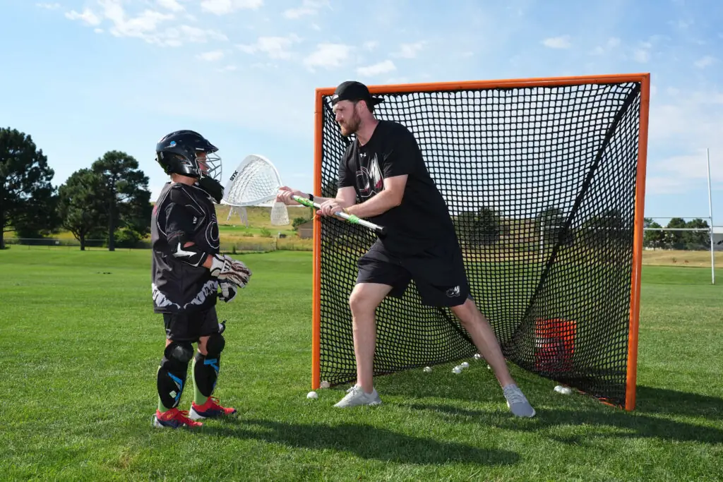 Adult coaching child in lacrosse goalkeeping on a sunny field.