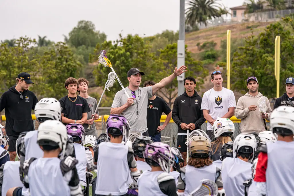 Coach in black cap giving instructions to gathered lacrosse players.