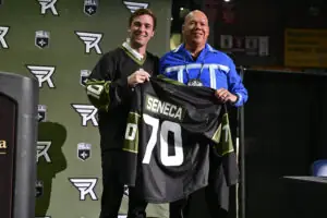Two men smiling and holding a jersey labeled "Seneca 70" at a sports event