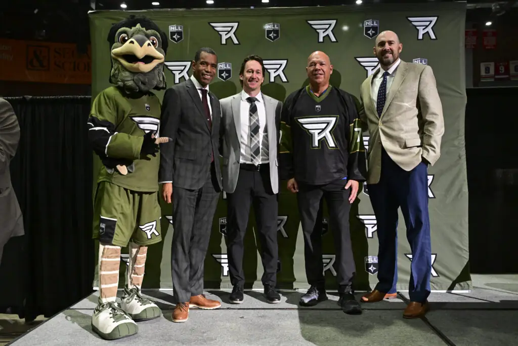 Mascot and four men pose in front of NLL backdrop, smiling.