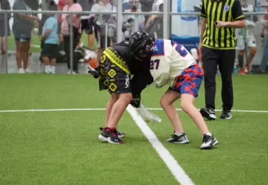 Two lacrosse players face off on an indoor field, watched by an official and audience