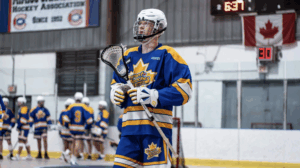 Liam-Aston playing lacrosse, wearing blue Toronto Beaches uniform, holding stick. Canadian flag and scoreboard in background