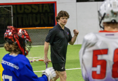 Instructor coaching lacrosse players indoors, gesturing with a fist. Players in blue and white jerseys facing the coach