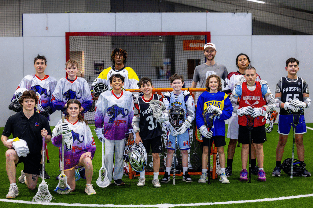 A group of young lacrosse players posing on an indoor field, wearing jerseys and protective gear, with coaches in the background.