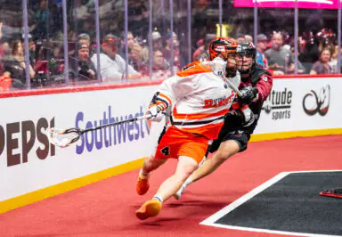 Buffalo Bandits player in white jersey and orange shorts, chased by Colorado Mammoth player, during lacrosse game