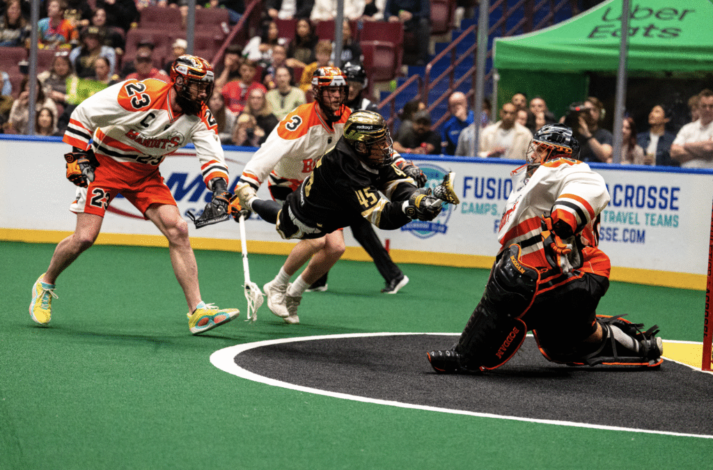 A lacrosse player in black is diving to shoot the ball while a goalie in orange prepares to block the shot, with teammates in the background.