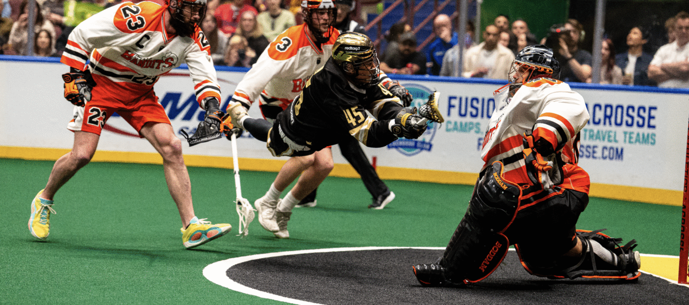 A lacrosse player in black is diving to shoot the ball while a goalie in orange prepares to block the shot, with teammates in the background.