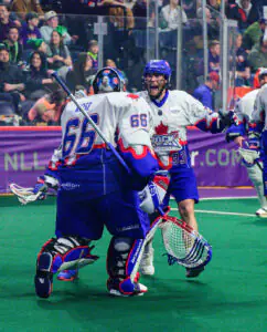 Two lacrosse players celebrating on the field, one wearing jersey 66 and the other 33, with a cheering crowd