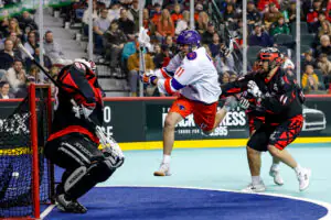 Player 1186 in white jumps and shoots at the goal during a lacrosse game, with defenders approaching