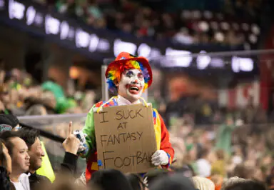 Clown in colorful costume holding sign saying "I suck at fantasy football" at a sports event