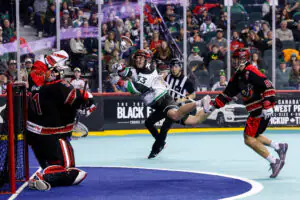 Player in white jersey attempts lacrosse goal against goalie in black, intense action with crowd watching