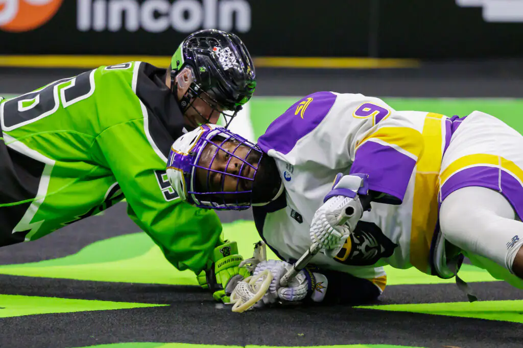 Lacrosse players in intense face-off, green and purple uniforms, on indoor field
