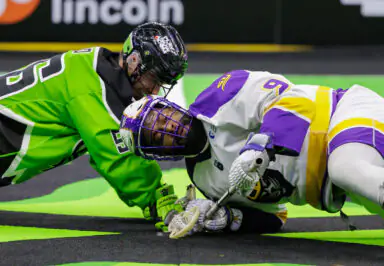 Lacrosse players in intense face-off, green and purple uniforms, on indoor field